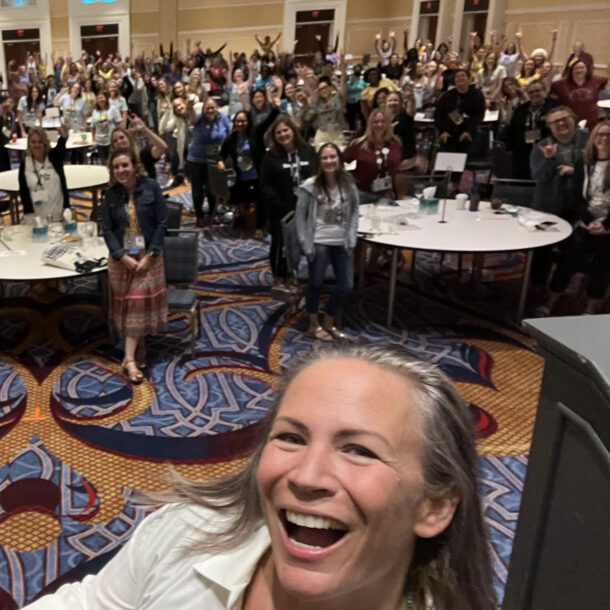 Rachel smiles in front of a large group of conference attendees who are cheering behind her.