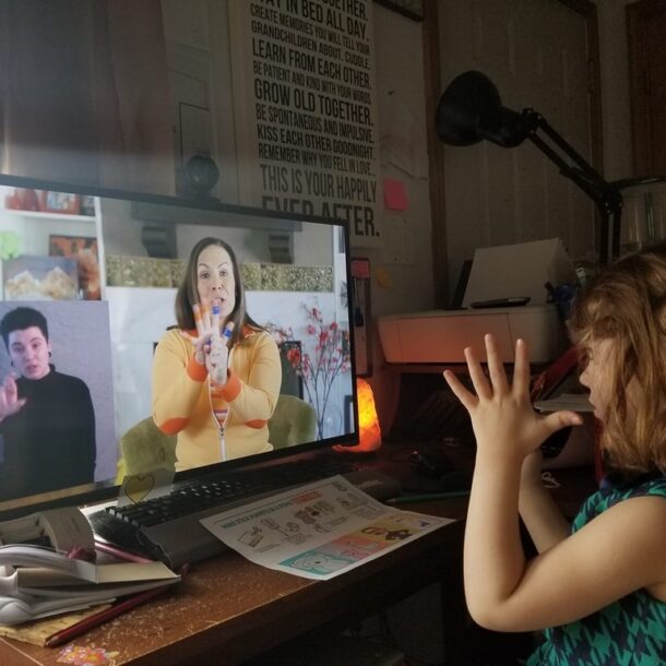 Rachel video chats with a little girl. The screen is split between Rachel and an ASL interpreter. A young child signs back to Rachel through the computer.